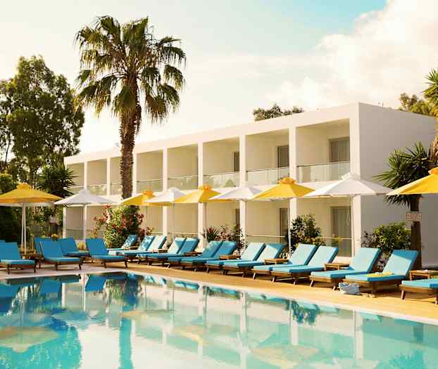 Et hotel med hvide bygninger og altaner i en grøn omgivelser, foran en pool med liggestole og parasoller. (AI-genereret tekst)