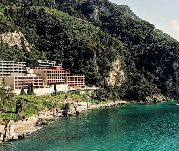 Hotel beliggende ved en klippefyldt kyst omgivet af frodige bjerge. (AI-genereret tekst)