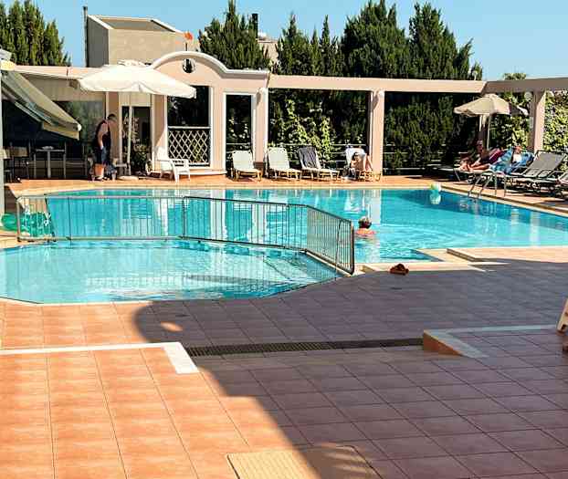 Udendørs swimmingpool med liggestole og parasoller ved siden, samt personer der svømmer og soler sig. (AI-genereret tekst)