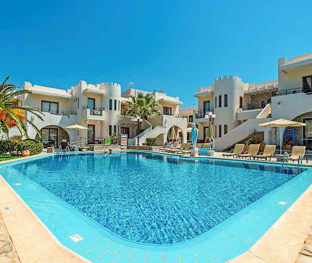 En swimmingpool omgivet af liggestole og parasoller foran bygninger med hvid facade og unikke arkitektoniske detaljer. (AI-genereret tekst)