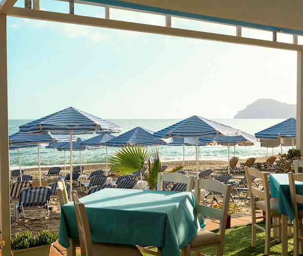 Strand med blå-hvidstribede parasoller og liggestole, borde i forgrunden. (AI-genereret tekst)