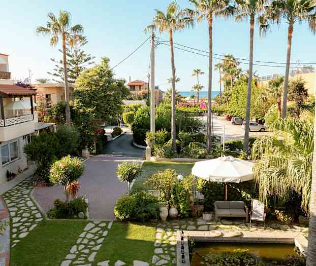 Billede af en have med en sofa og parasol nær en lille dam, omgivet af palmer og grønt. (AI-genereret tekst)