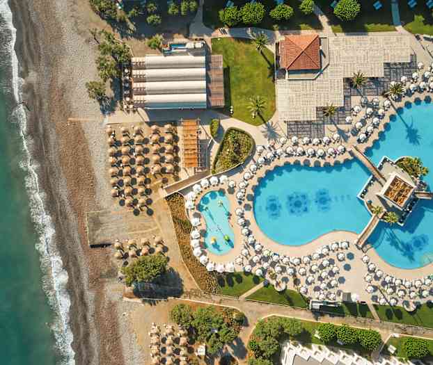 Luftfoto af en strand med parasoller og liggestole ved siden af et stort poolområde med blå bassiner og hvide parasoller. (AI-genereret tekst)
