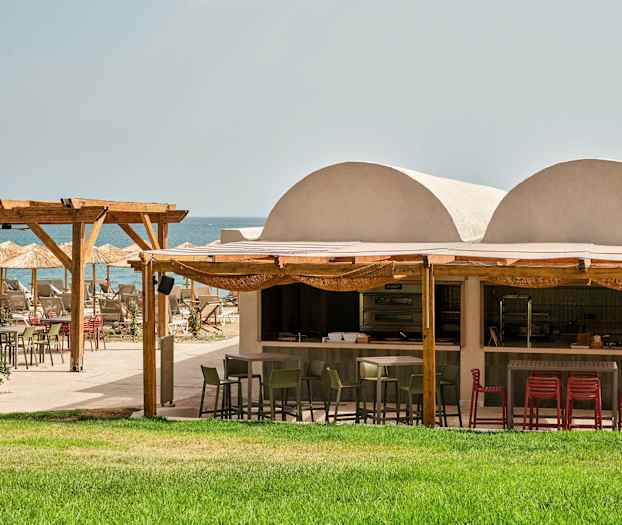 Udendørs restaurant ved stranden med grøn græsplæne, træpergola og parasoller. (AI-genereret tekst)