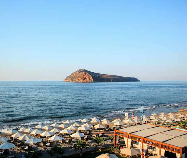 En strand med liggestole og parasoller ved havet, med en ø synlig i baggrunden. (AI-genereret tekst)