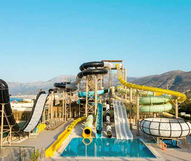 Vandpark med forskellige rutsjebaner og bassiner foran en bjergrig baggrund. (AI-genereret tekst)