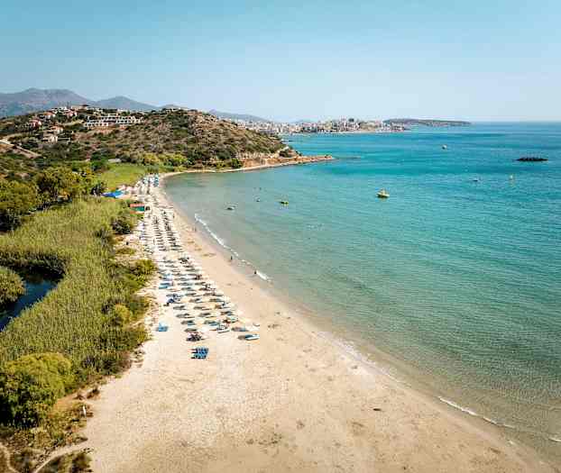 Sandstrand med solsenge ved havet, omgivet af grønne områder og bjerge i baggrunden. (AI-genereret tekst)