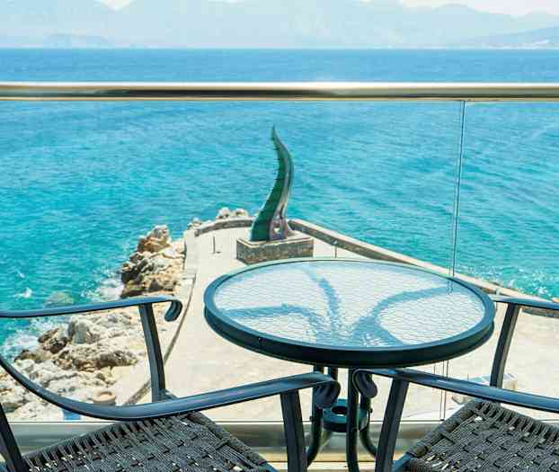 Udsigt fra en terrasse mod havet med to stole og et bord i forgrunden. (AI-genereret tekst)