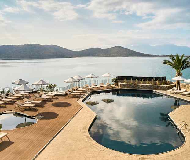 En pool med solsenge og parasoller ved havet, omgivet af bjerge og en palme. (AI-genereret tekst)