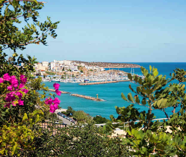 Billede af en havn med sejlbåde omgivet af bygninger, set gennem grønt og rosa blomster. (AI-genereret tekst)