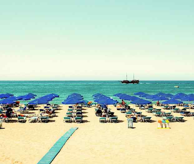 Strand med blå parasoller, liggestole og et skib på havet. (AI-genereret tekst)