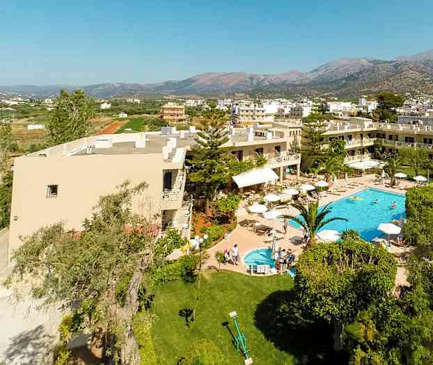 Luftfoto af hotel med pool og have, omgivet af grønne områder og bygninger med bjerge i baggrunden. (AI-genereret tekst)