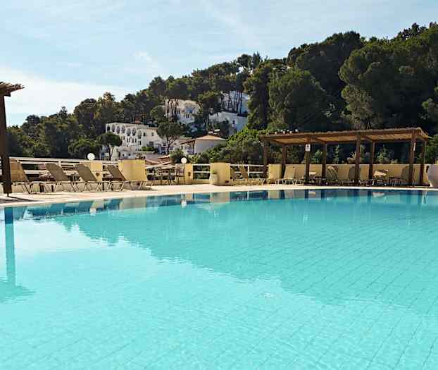 En udendørs swimmingpool omgivet af liggestole og pergola. I baggrunden er der træer og hvide bygninger på en skråning. (AI-genereret tekst)