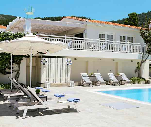 Hvid bygning med røde tage og poolområde med liggestole og parasoller. (AI-genereret tekst)