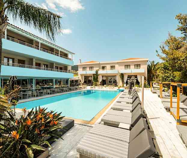 En hotelpool omgivet af liggestole og bygninger under en blå himmel. (AI-genereret tekst)