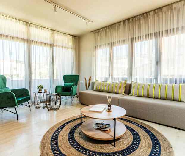 Et lyst opholdsrum med store vinduer, to grønne lænestole, en beige sofa med stribede puder og et rundt sofabord med dekorationer. (AI-genereret tekst)