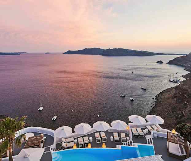 En udsigt over en kystlinje med havet ved solnedgang. I forgrunden ses en pool og hvide liggestole med parasoller. (AI-genereret tekst)