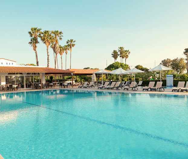 Udendørs swimmingpool med liggestole og parasoller på siden. Bagved ses en bygning med skiltet 'SNACK BAR' og palmer i baggrunden. (AI-genereret tekst)