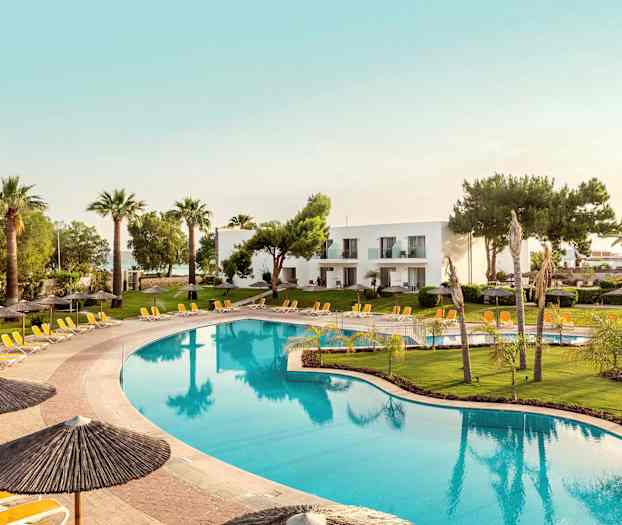 Stor pool omkranset af liggestole og parasoller, med grønne græsplæner og palmer i baggrunden samt hvide bygninger. (AI-genereret tekst)