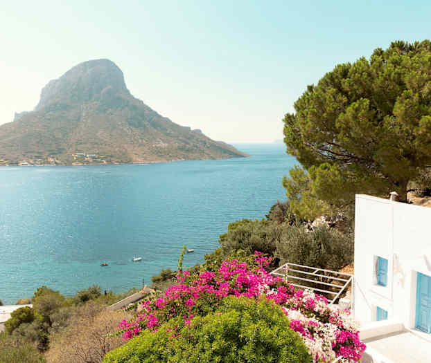 En hvid bygning med blå døre, omgivet af grønt løv og lyserøde blomster, med udsigt over havet og et bjerg i baggrunden. (AI-genereret tekst)