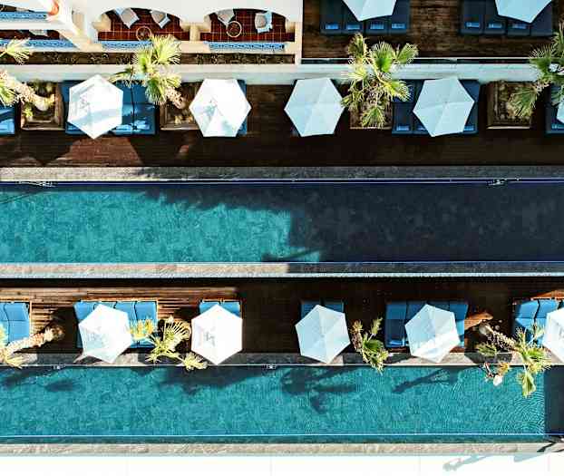 Et fugleperspektivbillede af to parallelle swimmingpools omgivet af palmer, liggestole og parasoller. (AI-genereret tekst)