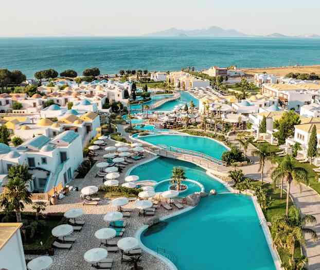 Udsigt over en ferieby med swimmingpools, hvide bygninger og havudsigt i baggrunden. (AI-genereret tekst)