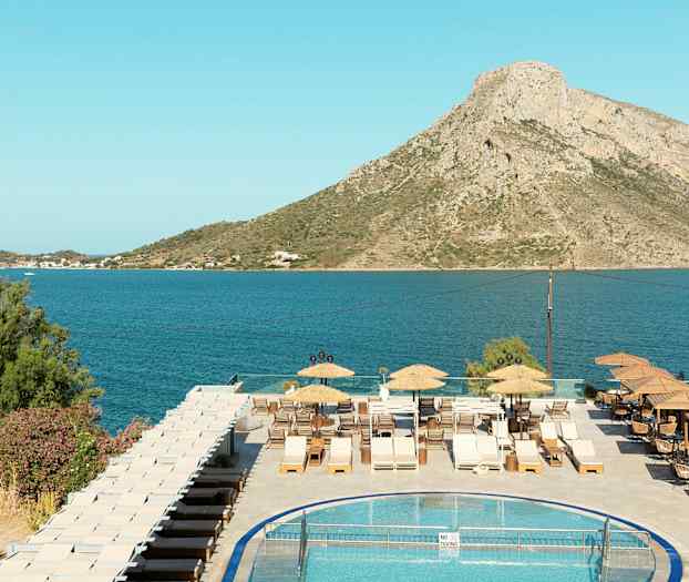 Udsigt over en pool, en terrasse med solvogne, og en havudsigt med bjerge i baggrunden. (AI-genereret tekst)