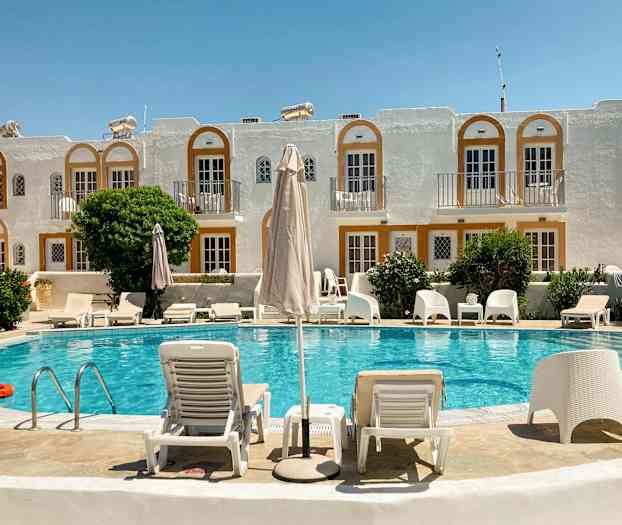 En hotelpool med liggestole og parasoller foran en bygning med hvide facader og orange detaljer. (AI-genereret tekst)