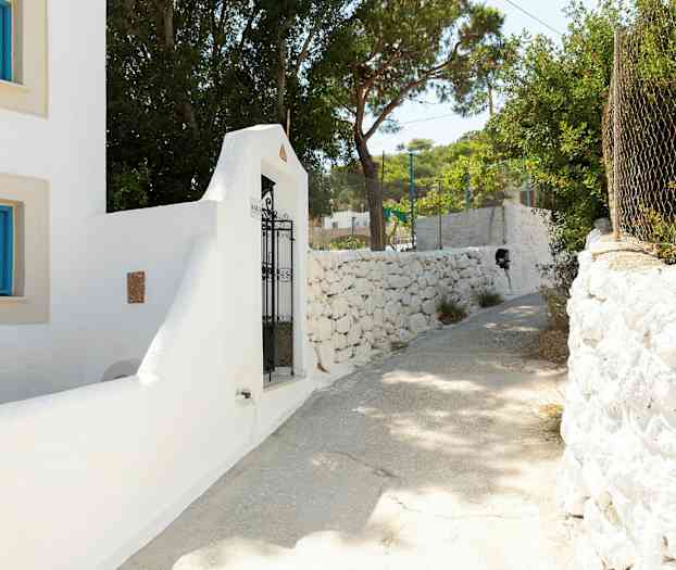 Hvid bygning med blå vinduesrammer og en jernlåge ved en stenmur. Solrig gangsti omgivet af træer. (AI-genereret tekst)