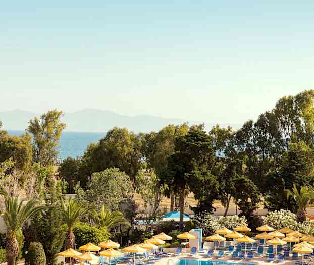 Poolområde med gule parasoller omgivet af grønne områder og udsigt til havet. (AI-genereret tekst)