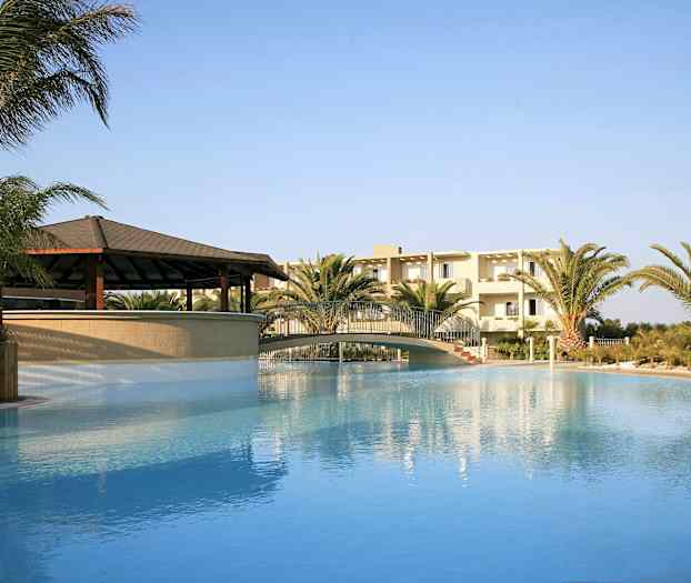 Udendørs swimmingpool med bro, omgivet af palmer og en bygning i baggrunden. (AI-genereret tekst)