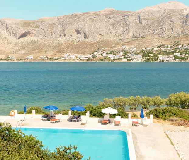 Udsigt over en pool med liggestole og parasoller, med havet og bjerge i baggrunden. (AI-genereret tekst)