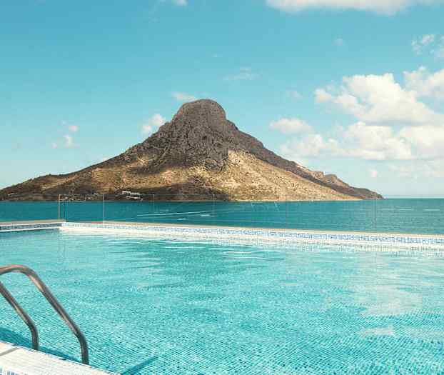 En pool med klart blåt vand foran et bjerg ved havet under en klar himmel. (AI-genereret tekst)