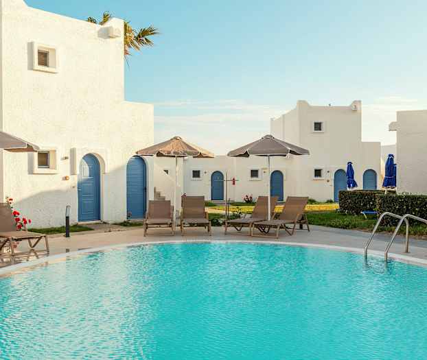 En pool omgivet af liggestole og parasoller, med hvide bygninger med blå døre i baggrunden. (AI-genereret tekst)