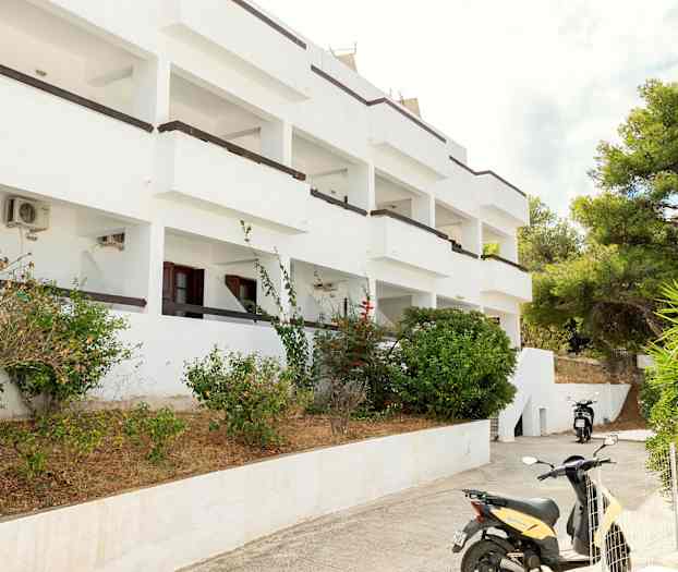 Hvid bygning med altaner omgivet af buske og en parkeret motorcykel. (AI-genereret tekst)