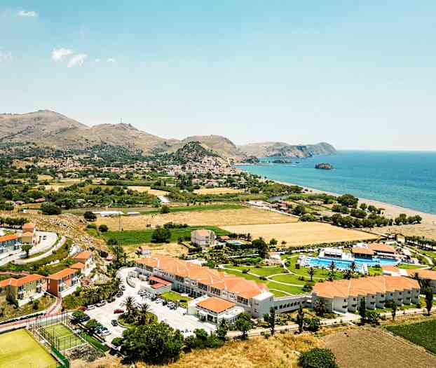 Luftfoto af et kystlandskab med hotelbygninger, grønne områder og havet i baggrunden. (AI-genereret tekst)