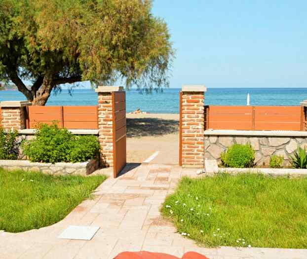 En grøn have med en stenbelagt gangsti fører til en strand bag et træhegn med en åben port. (AI-genereret tekst)