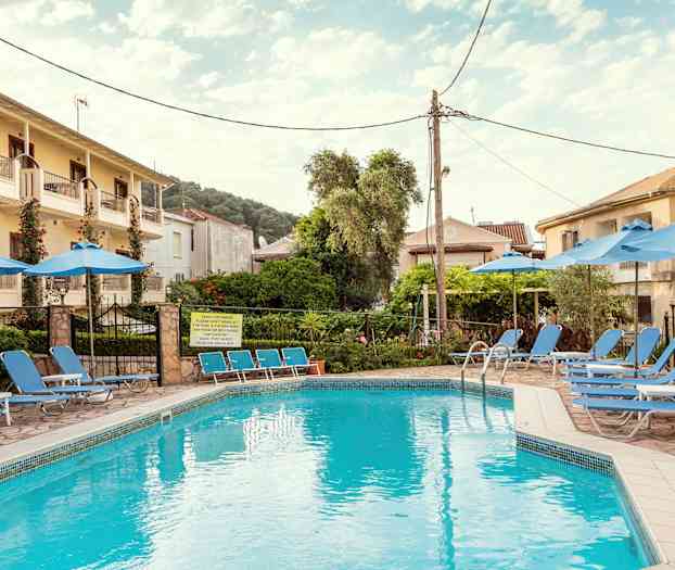 En pool omgivet af liggestole og parasoller, med bygninger og træer i baggrunden. (AI-genereret tekst)