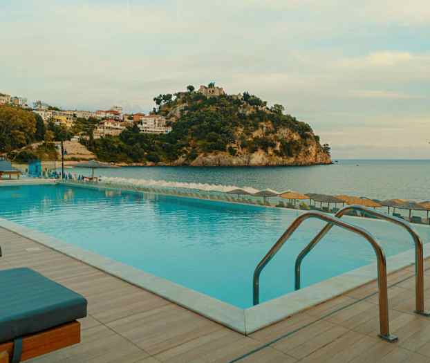 En pool med liggestole og parasoller ved havet med en bjergrig kystlinje i baggrunden. (AI-genereret tekst)