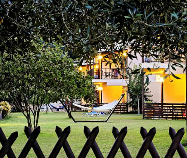 Billede af en have med græsplæne, hængekøje og omgivende vegetation. I baggrunden ses et hus med oplyste altaner. (AI-genereret tekst)