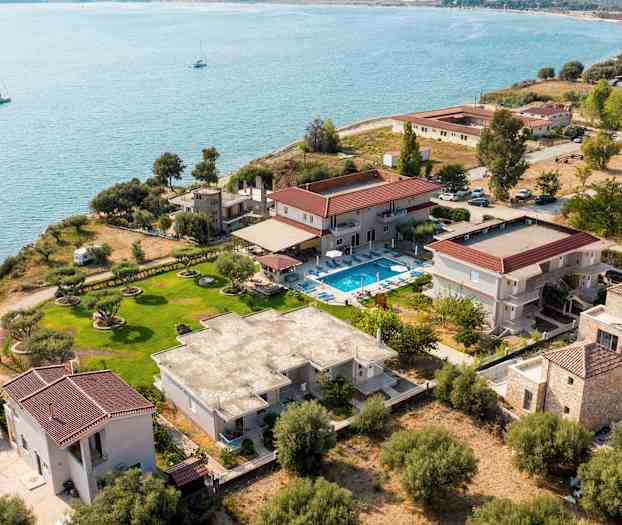 Fugleperspektiv af en kystejendom med forskellige bygninger, en græsplæne og en pool tæt på vandet. (AI-genereret tekst)