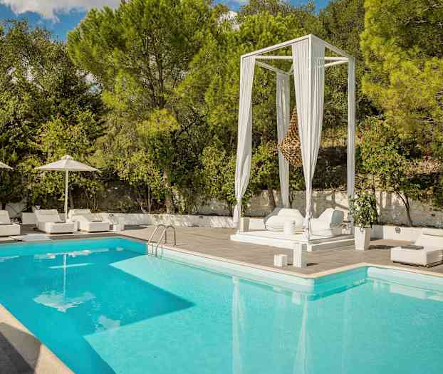 Swimmingpool med omkringliggende hvide liggestole og parasoller samt frodige træer i baggrunden. (AI-genereret tekst)