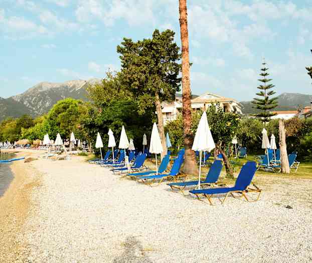 En strand med liggestole og parasoller, omgivet af grønt område og bygninger i baggrunden. (AI-genereret tekst)