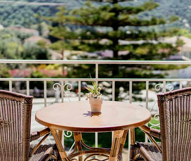 En altan med et træbord og to rattanstole samt en lille blomsterpotte med blomster på bordet. Træer og bygninger ses i baggrunden. (AI-genereret tekst)