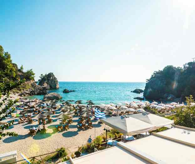 Sandstrand med parasoller og solsenge ved det klare blå hav, omgivet af klipper og grønt. (AI-genereret tekst)