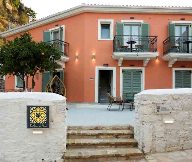 En lille hotelbygning med orange facade og grønne skodder samt altaner. Foran bygningen er en siddegruppe og et træ med frugt. (AI-genereret tekst)