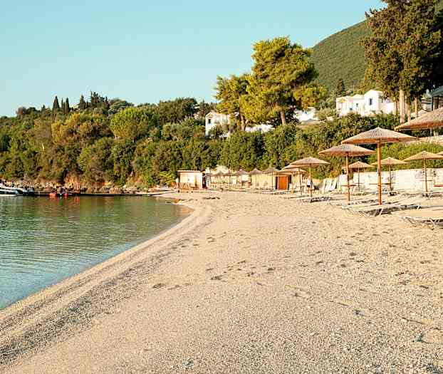 En strand med liggestole og parasoller, vegetation og bygninger i baggrunden. (AI-genereret tekst)