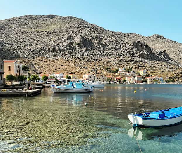 En havn med klart vand, små både og farverige huse ved foden af stenede bjerge. (AI-genereret tekst)