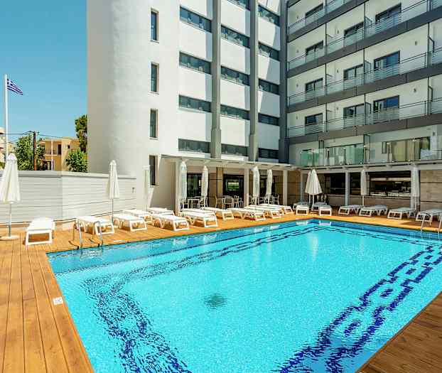 Hotelpool med liggestole og parasoller, omgivet af bygninger. (AI-genereret tekst)