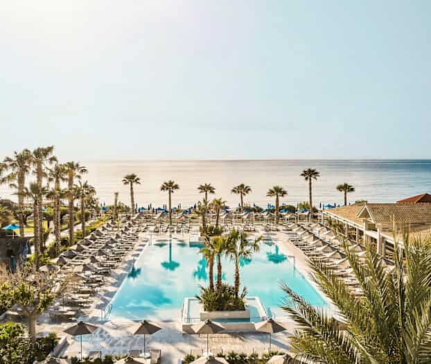 En stor udendørs pool omgivet af liggestole og parasoller med havet i baggrunden. (AI-genereret tekst)